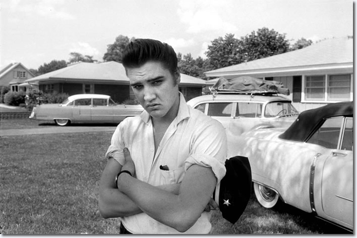 Elvis in the front yard of his home at 1034 Audubon Drive in May 1956. When this photo was taken, Elvis had just returned from touring; the band’s instruments were still packed on the roof of his car. His famous pink Cadillac can be seen over his right shoulder,