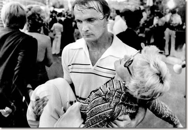 An exhausted volunteer carries a victim of the heat and confusion to a first aid station inside the gates of Graceland Mansion in Memphis, where thousands gathered for a chance to view Elvis Presley's body. 